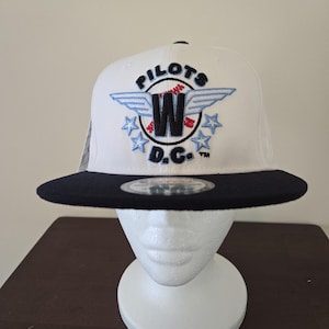 Peut inclure: Casquette de baseball blanche et noire avec le texte "PILOTS W D.C." brodé en bleu, rouge et noir. La casquette a une visière plate et une couronne structurée. Le chapeau est présenté sur une tête de mannequin blanche.