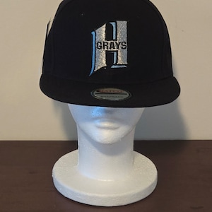 Peut inclure: Une casquette de baseball noire et blanche avec un "H" blanc et le mot "GRAYS" brodé sur le devant. La casquette a une fermeture à pression noire.