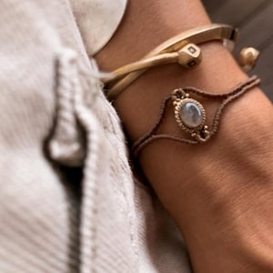 Op de afbeelding: Een close-up van een vrouwenpols met drie armbanden. De bovenste armband is een zilveren lepel die tot een cirkel is gebogen. De middelste armband is een gouden armband met een vierkant ontwerp. De onderste armband is een bruin koord met een kleine ovale stenen hanger.