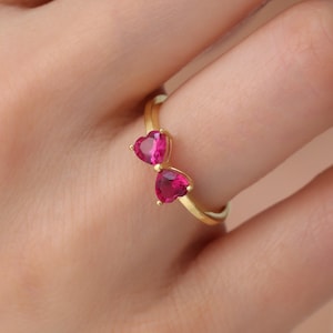 Puede incluir: Un anillo de oro con dos piedras preciosas en forma de corazón rosa. El anillo está en el dedo de una persona.