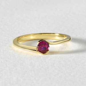 May include: A gold ring with a vibrant red gemstone. The ring features a slender, curved band that gracefully wraps around the finger, culminating in a bezel setting that holds the oval-shaped ruby. The ring is set against a white background.