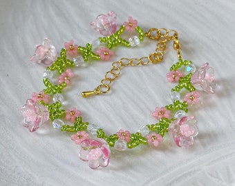May include: A delicate bracelet made with pink and green glass beads, featuring small flower shapes. The bracelet has a gold chain closure.