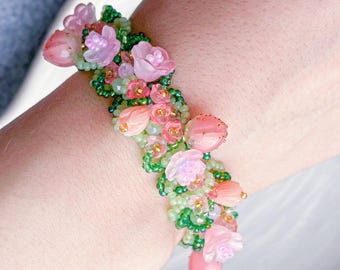 May include: A floral bracelet featuring pink, peach, and green beads and flower accents. The bracelet is worn on a wrist and is set against a light background. The bracelet's design includes small, delicate flowers and beads.