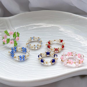 May include: Six beaded rings with flower designs. The rings are in various colors, including pink, blue, green, red, and white.