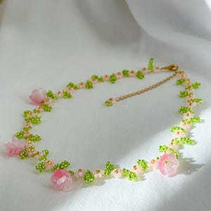 May include: A delicate pink and green beaded necklace with small flower accents. The necklace is made with tiny glass beads and features a gold chain closure.