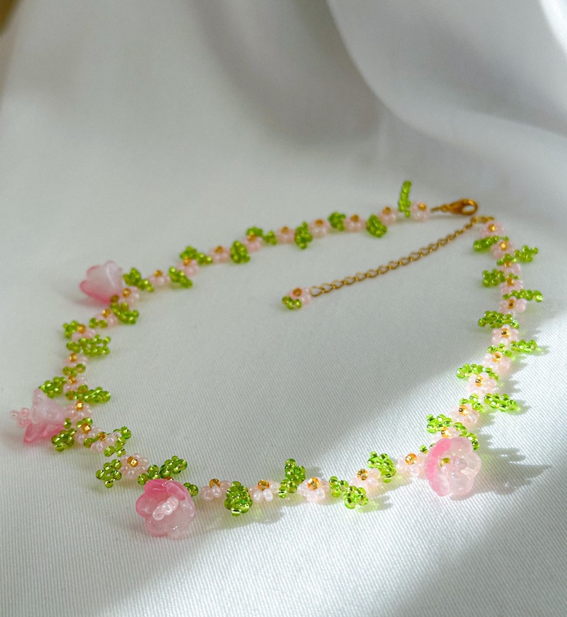 May include: A delicate pink and green beaded necklace with small flower accents. The necklace is made with tiny glass beads and features a gold chain closure.