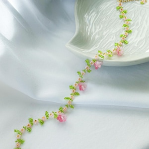 May include: A delicate beaded necklace with pink flowers and green leaves. The necklace is made of small beads and features a floral design.