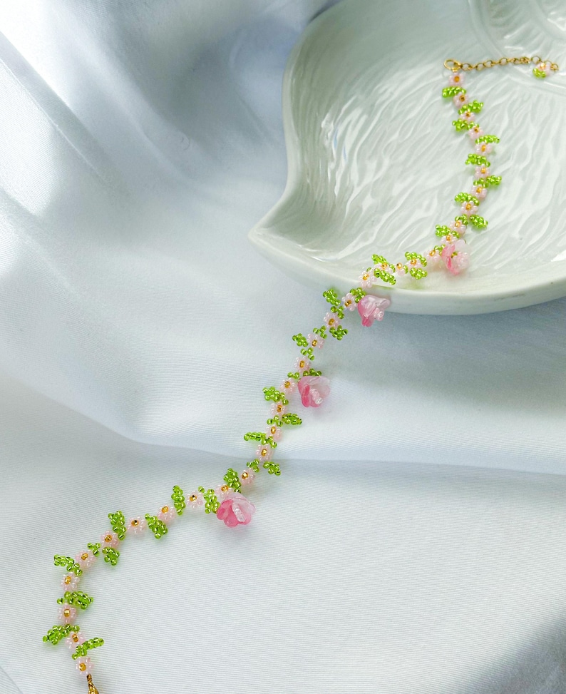 May include: A delicate beaded necklace with pink flowers and green leaves. The necklace is made of small beads and features a floral design.