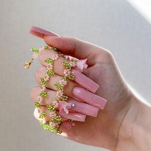 May include: A delicate pink and green beaded necklace with small flower charms. The necklace is held in a hand with long, pink acrylic nails.