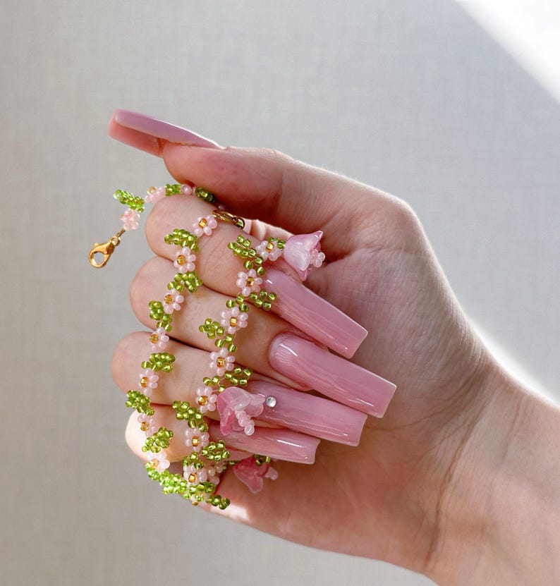 May include: A delicate pink and green beaded necklace with small flower charms. The necklace is held in a hand with long, pink acrylic nails.