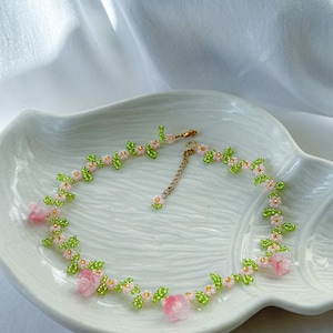 May include: A delicate beaded necklace with pink flowers and green leaves. The necklace is made with small beads and has a gold chain clasp.