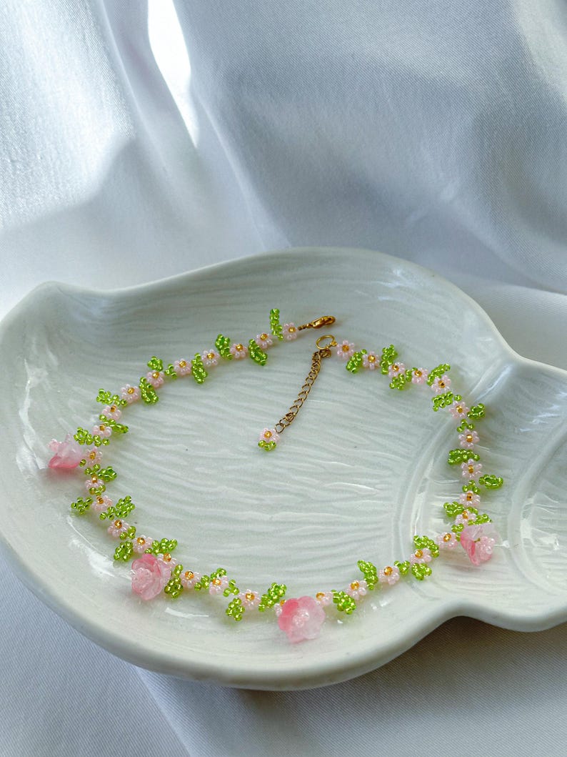 May include: A delicate beaded necklace with pink flowers and green leaves. The necklace is made with small beads and has a gold chain clasp.