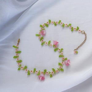May include: A delicate beaded necklace with pink flowers and green leaves. The necklace is made of small beads and features a gold chain clasp.
