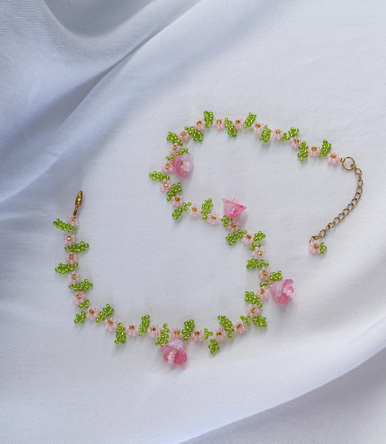 May include: A delicate beaded necklace with pink flowers and green leaves. The necklace is made of small beads and features a gold chain clasp.