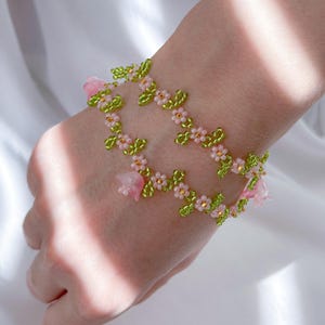 May include: A delicate bracelet made with pink and green beads, featuring small flower and leaf designs. The bracelet is made with a string of beads and has a floral theme.