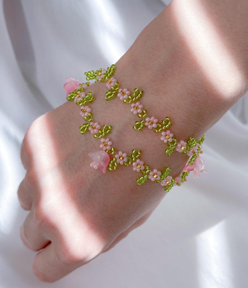 May include: A delicate bracelet made with pink and green beads, featuring small flower and leaf designs. The bracelet is made with a string of beads and has a floral theme.