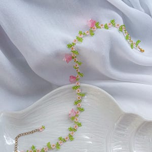 May include: A delicate beaded necklace with pink flowers and green leaves. The necklace is made of small beads and has a gold chain closure.