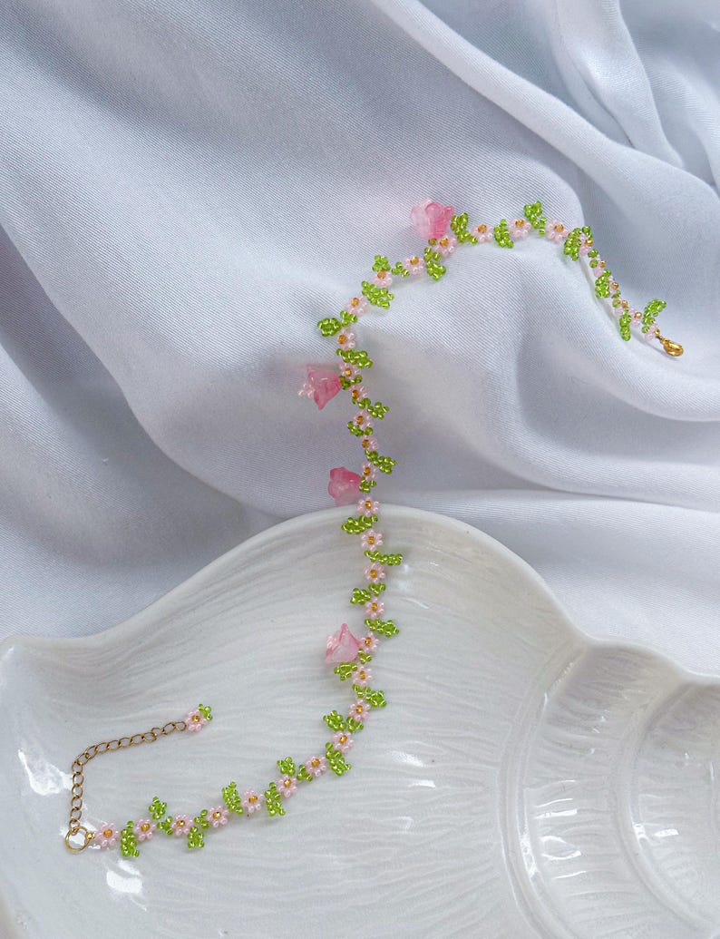 May include: A delicate beaded necklace with pink flowers and green leaves. The necklace is made of small beads and has a gold chain closure.