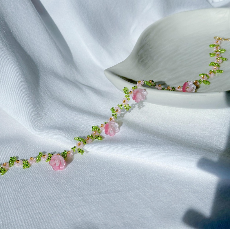 May include: A delicate beaded necklace with pink and white flower accents and green leaves. The necklace is made of small beads and is strung on a thin wire.