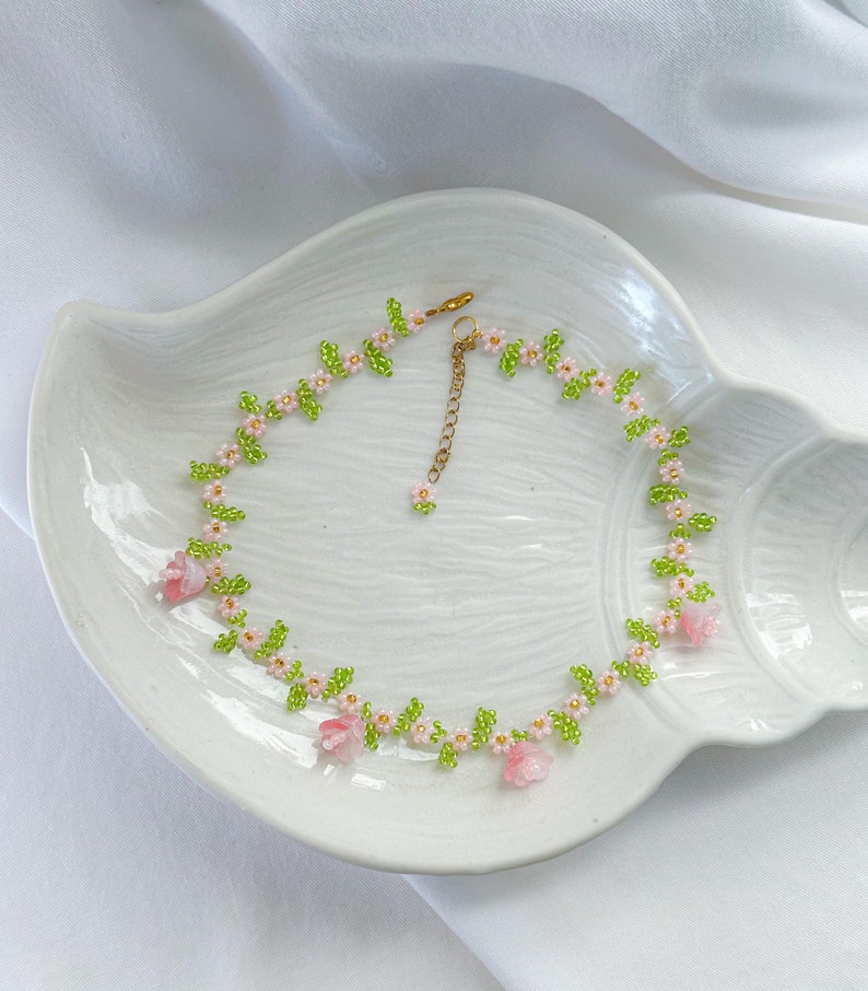 May include: A delicate floral necklace made with pink and green beads. The necklace features small pink flowers and green leaves, creating a dainty and feminine look.