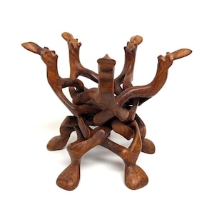 May include: A handcrafted wooden decorative stand with a unique, intertwined design. The stand is made of dark brown wood and features multiple curved arms and legs, creating an artistic and functional piece. The stand is designed to hold a bowl or other decorative items.
