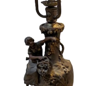 May include: A bronze-toned metal candelabra lamp featuring two light sockets with leaf-shaped bobeches. The central column is adorned with a sculpted figure of a person holding a basket of fruit, set against a textured vase-shaped base.