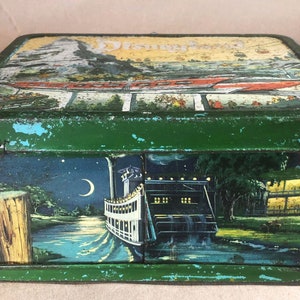 Puede incluir: Fiambrera vintage de metal verde con una escena de Disneyland en la tapa y una escena nocturna de un barco de vapor en el lateral. La tapa presenta una montaña con un teleférico y el texto "Disneyland".