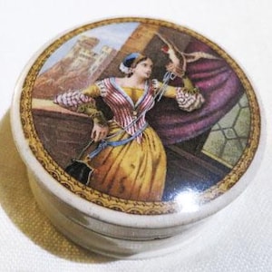May include: A round porcelain trinket box with a lid featuring a colorful illustration of a woman in a yellow dress and blue hat standing on a balcony with a bird in her hand. The background shows a castle and a red curtain.