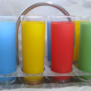 Puede incluir: Un juego de seis vasos altos y transparentes llenos de líquidos de colores azul, amarillo, rojo y verde. Los vasos están colocados en un soporte metálico plateado con un asa curvada, ideal para servir bebidas.