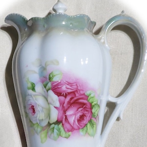 Puede incluir: Una tetera de porcelana blanca con una tapa verde y un diseño floral con rosas rosas y blancas. La tetera tiene un asa y un pico.
