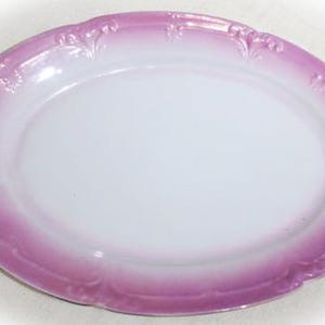 May include: An oval-shaped ceramic serving platter featuring a smooth white center and a wide, graduated pink border. The rim is decorated with a subtle, raised embossed pattern along the edge, providing a textured finish to the plate.