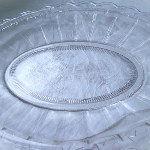 May include: A clear, oval-shaped glass serving platter with a scalloped edge. The platter features a textured design and a raised, oval center. The overall design is simple and elegant, suitable for serving food.