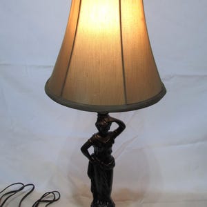 May include: A table lamp featuring a dark brown, sculpted base in the shape of a standing figure with one hand raised to the head. The lamp is topped with a tapered, light tan fabric shade and a dark finial, connected to a black electrical cord.