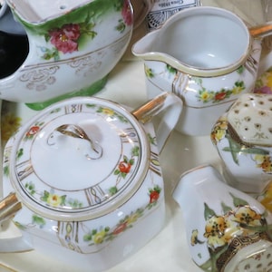 Puede incluir: Un juego de tazas y platillos de té de porcelana vintage con motivos florales. Las tazas y los platillos son blancos con ribetes dorados y presentan delicados diseños florales en tonos rosa, amarillo y verde.
