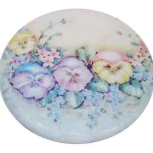 May include: A round ceramic plate with a painted design of three pansies in shades of yellow, pink, and purple. The pansies are surrounded by smaller blue flowers and green leaves.