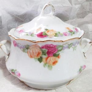Puede incluir: Fuente de porcelana blanca con tapa y diseño floral de rosas en tonos rosa, melocotón y amarillo. La fuente tiene ribetes y asas doradas, y una tapa a juego con un asa de bucle.