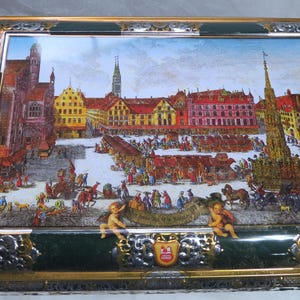 May include: A rectangular decorative box featuring a detailed illustration of a European town square, with buildings, people, and horse-drawn carriages. The box has a green and gold frame with ornate silver accents and cherub figures.