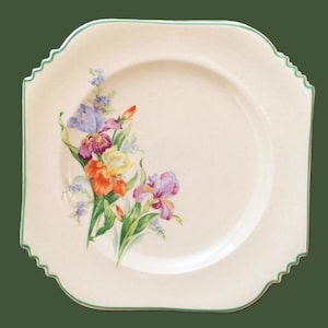 May include: A square, off-white plate with a scalloped edge and a green trim. The plate features a watercolor floral design of irises in shades of purple, orange, yellow, and blue. The flowers are clustered on the left side of the plate.