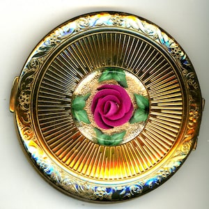 May include: A round, gold-toned compact with a central design featuring a pink rose surrounded by green leaves. The compact has a sunburst pattern radiating from the center and ornate floral detailing around the edge.