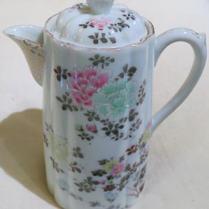以下が含まれることがあります： ピンク、緑、黄色で花柄が描かれた、蓋付きの白い磁器製のクリーマー。クリーマーは金色の縁取りが施された注ぎ口とハンドル、そして蓋のつまみに装飾的な金のアクセントが付いています。