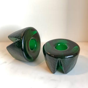 Puede incluir: Dos candelabros de vidrio verde oscuro con superficies texturizadas. Uno tiene forma redondeada con una abertura circular, y el otro tiene un diseño angular único. Los candelabros se exhiben sobre una superficie blanca.