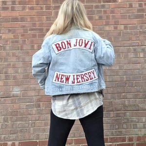 Puede incluir: Chaqueta vaquera azul claro con las palabras "BON JOVI" y "NEW JERSEY" en letras rojas. La chaqueta se lleva sobre una camisa a cuadros y leggings negros. El fondo es una pared de ladrillo rojo.