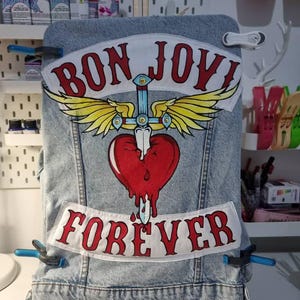 Op de afbeelding: Een lichtblauwe spijkerjas met een grote patch met de woorden "BON JOVI" boven een hart doorboord door een zwaard, met vleugels, en het woord "FOREVER" eronder. De patches zijn wit met rode letters en accenten.