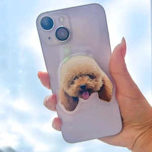 May include: A white iPhone case with a clear sticker of a brown and white poodle with its tongue sticking out.