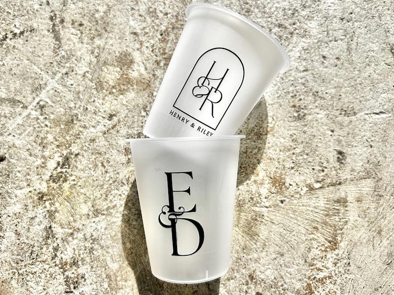Może przedstawiać: Dwa białe plastikowe kubki z czarnym nadrukiem. G&oacute;rny kubek ma napis "Henry & Riley" i wz&oacute;r monogramu. Dolny kubek ma napis "E & D" i wz&oacute;r monogramu.