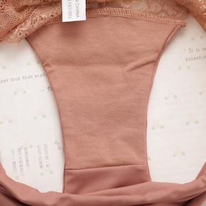 May include: A close-up of the inside of a pair of underwear. The underwear is made of a soft, peach-colored fabric. The fabric is smooth and has a slight sheen. The underwear has a lace trim around the top edge. The tag on the underwear reads "(165/85) CHINA".