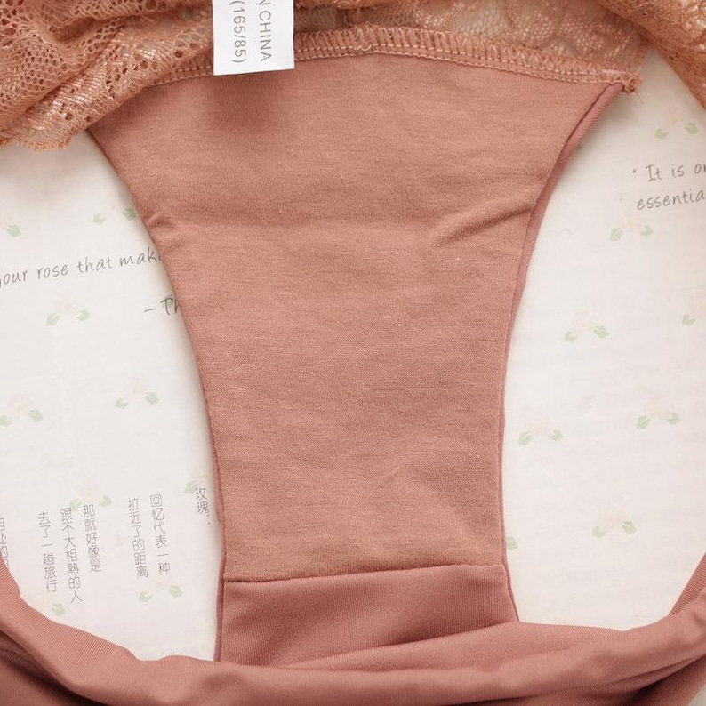 May include: A close-up of the inside of a pair of underwear. The underwear is made of a soft, peach-colored fabric. The fabric is smooth and has a slight sheen. The underwear has a lace trim around the top edge. The tag on the underwear reads "(165/85) CHINA".