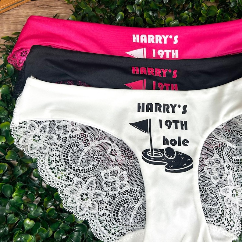May include: White lace underwear with the text "HARRY'S 19TH hole" printed on the front. The underwear is layered on top of two other pairs of underwear, one black and one pink, with the text "HARRY'S 19TH" printed on each.
