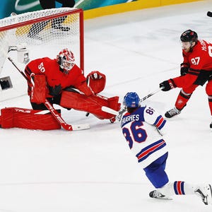 May include: A hockey goalie in a red uniform with a black maple leaf logo defends a net against two opposing players. One player wears a white jersey with blue and red accents and the number 86, while the other wears a red jersey with the number 97.
