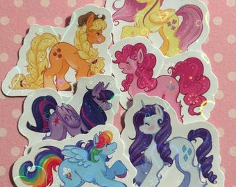 May include: A collection of six holographic stickers featuring stylized ponies in various colors including yellow, orange, pink, purple, blue, and white, each with unique manes, tails, and distinct cutie marks on their flanks.